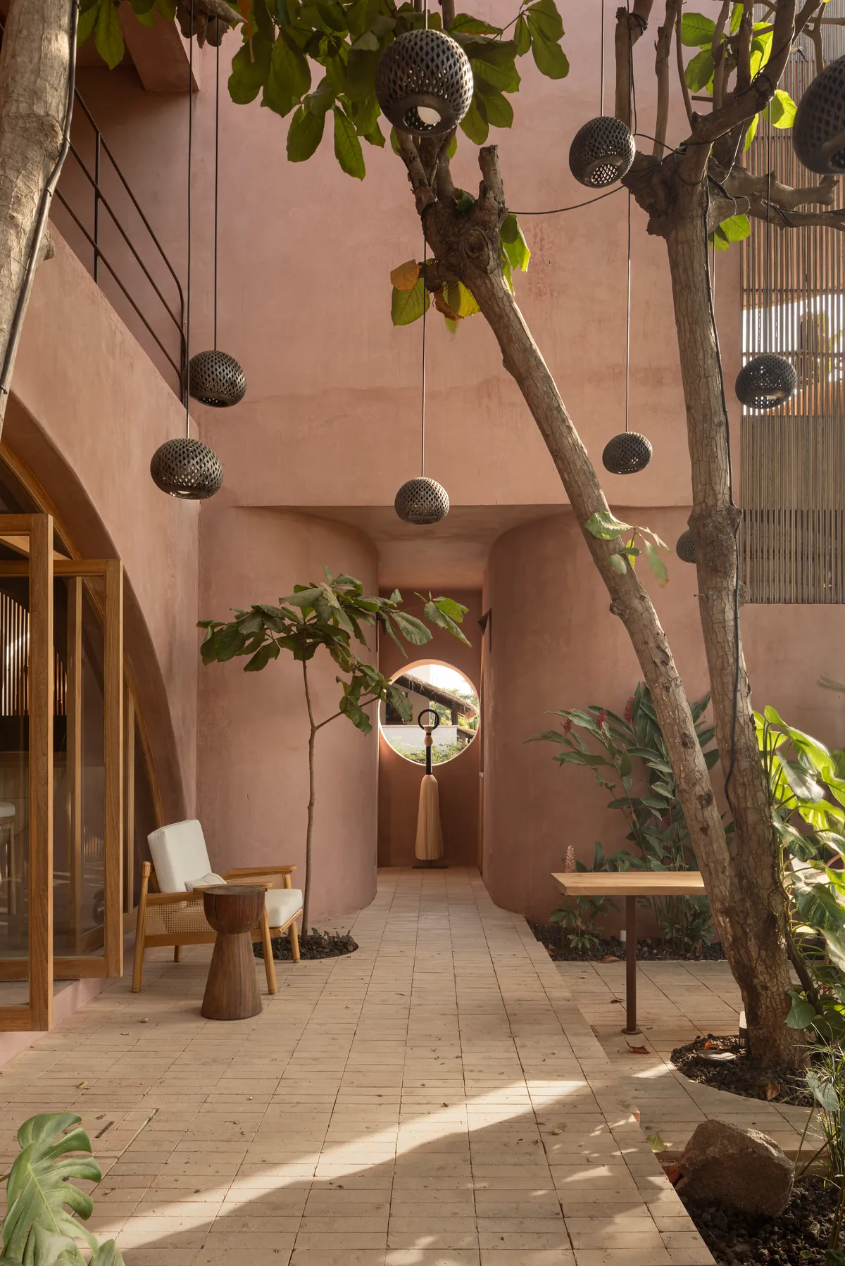 Patio interior con muros curvos de chukum rosado, árboles que crecen entre la arquitectura, lámparas de cerámica calada y una ventana circular al fondo