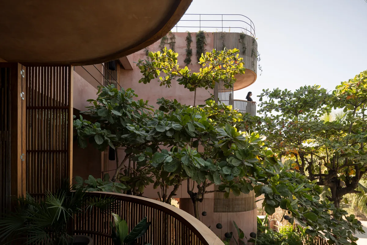 Desde la terraza de una suite Grand Signature: volúmenes curvos de chukum rosado con celosías y enredaderas entre árboles; una mujer en un balcón superior