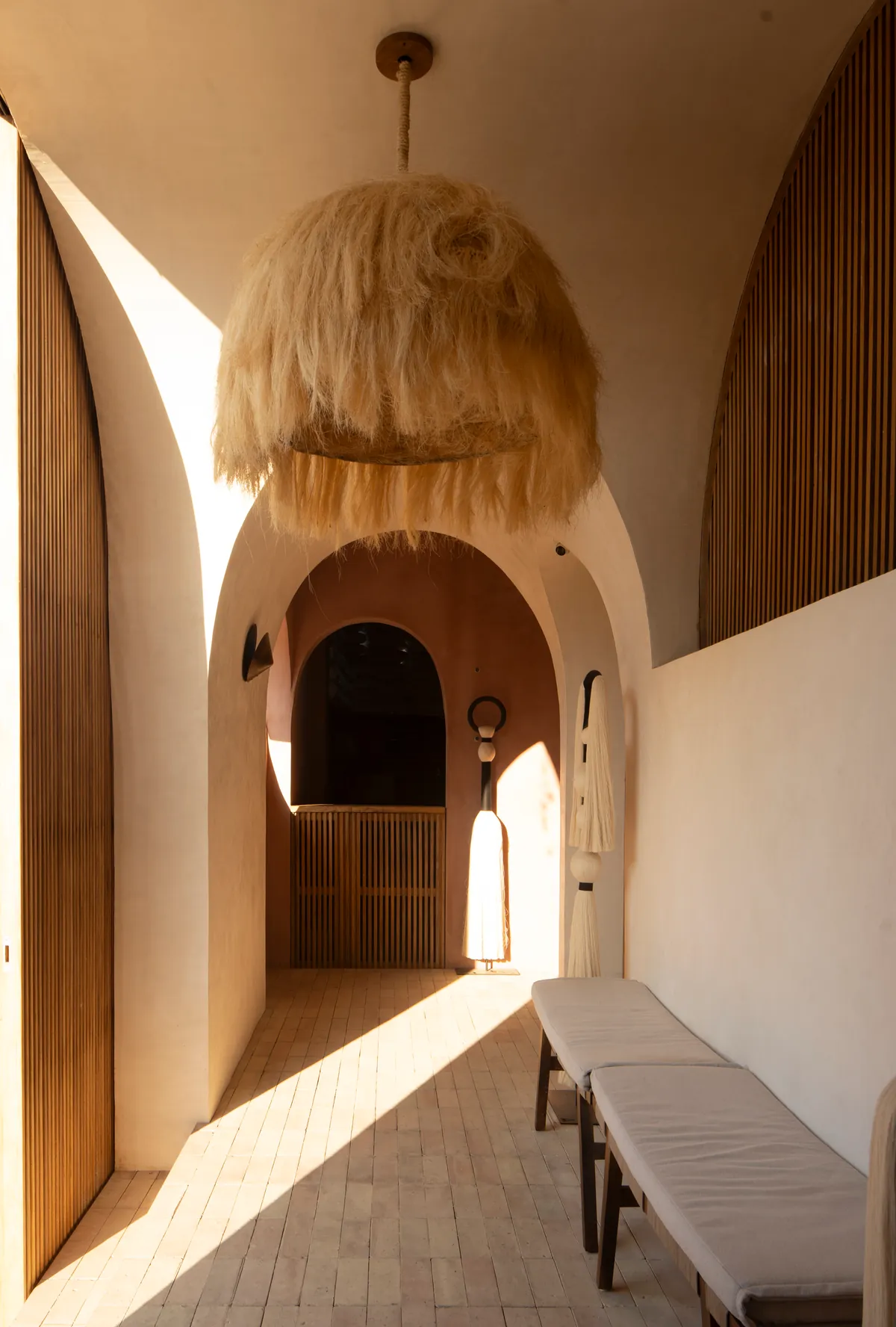 Galería de entrada con arcos encadenados, puertas altas de celosía de madera, una gran lámpara de fibra natural y bancas sobre piso de ladrillo cruzado por una franja de sol