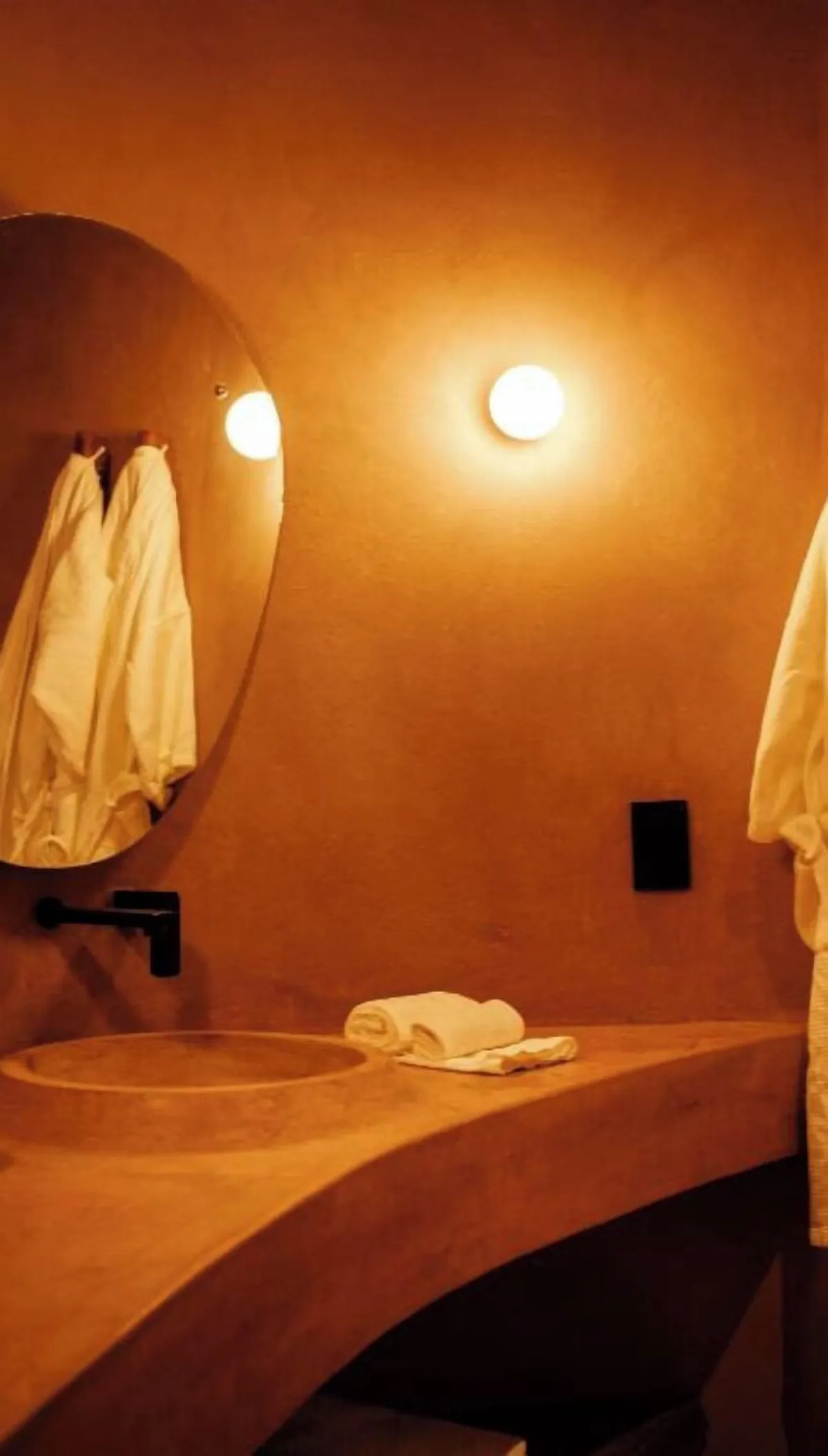 Pacific Terrace: lavabo de chukum con espejo redondo, batas blancas colgadas y luz cálida de aplique