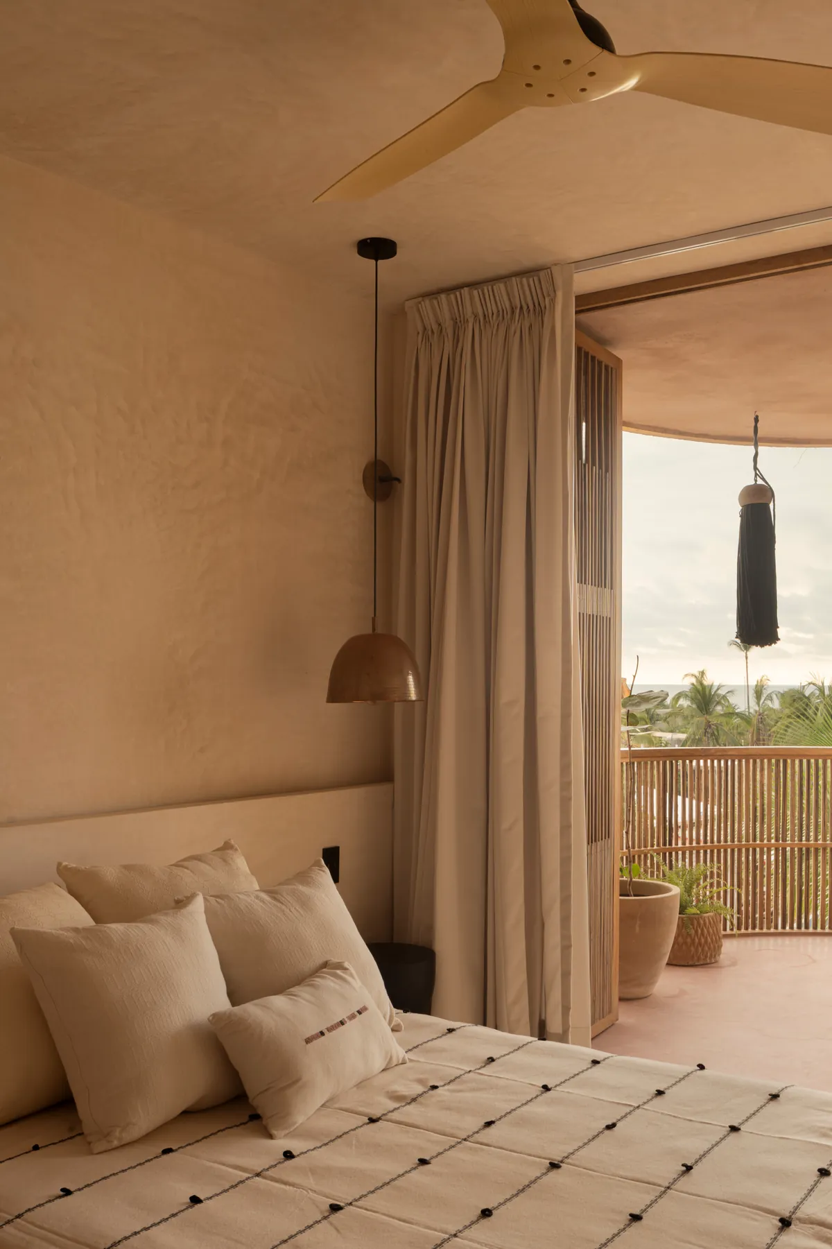 Pacific Terrace: cama con cojines claros junto a la puerta abierta al balcón; al fondo, palmeras y una franja de mar entre la bruma