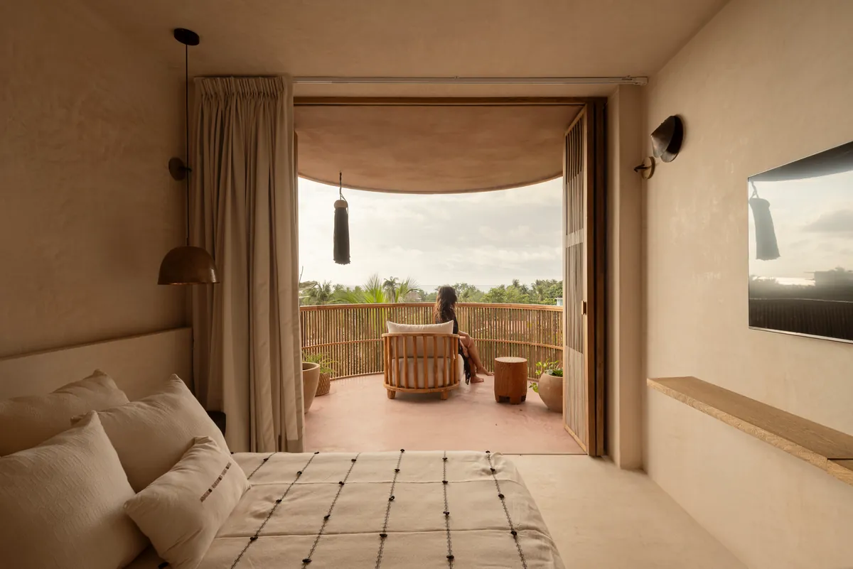 Suite Pacific Terrace: puertas de madera abiertas a un balcón curvo con barandal de varas, una cortina color arena y una mujer que mira las palmeras
