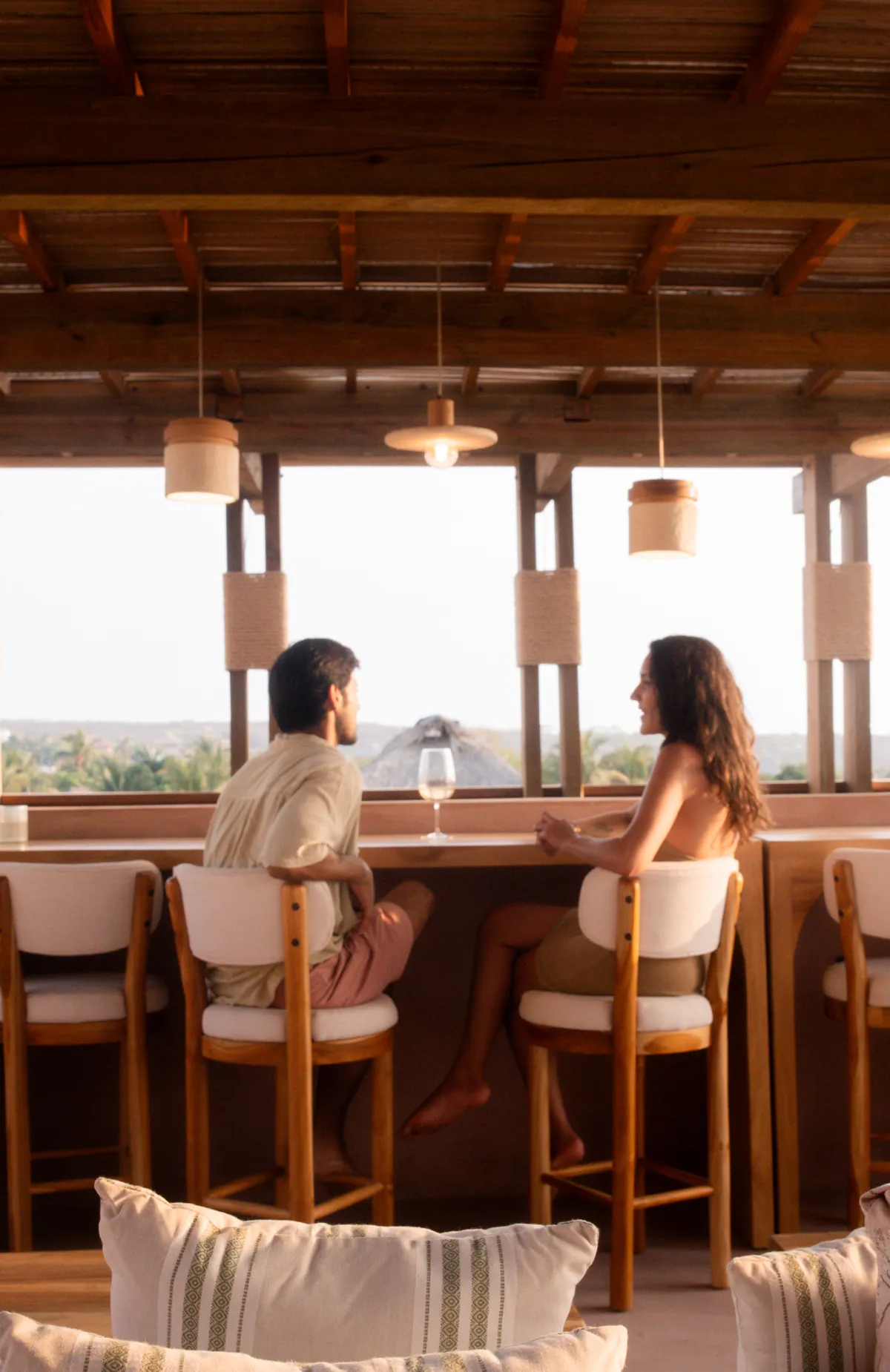 Una pareja conversa en la barra de madera de la azotea, frente a vanos abiertos con copas de palmeras y un techo de palapa