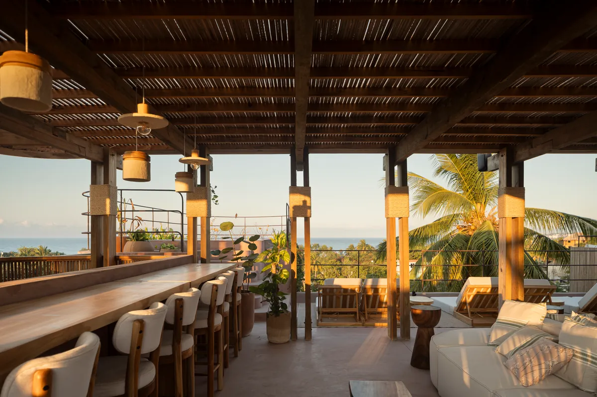 Azotea bajo una pérgola de madera con lámparas colgantes, una barra larga de madera con bancos altos, camastros y palmeras; al fondo, una franja del Pacífico