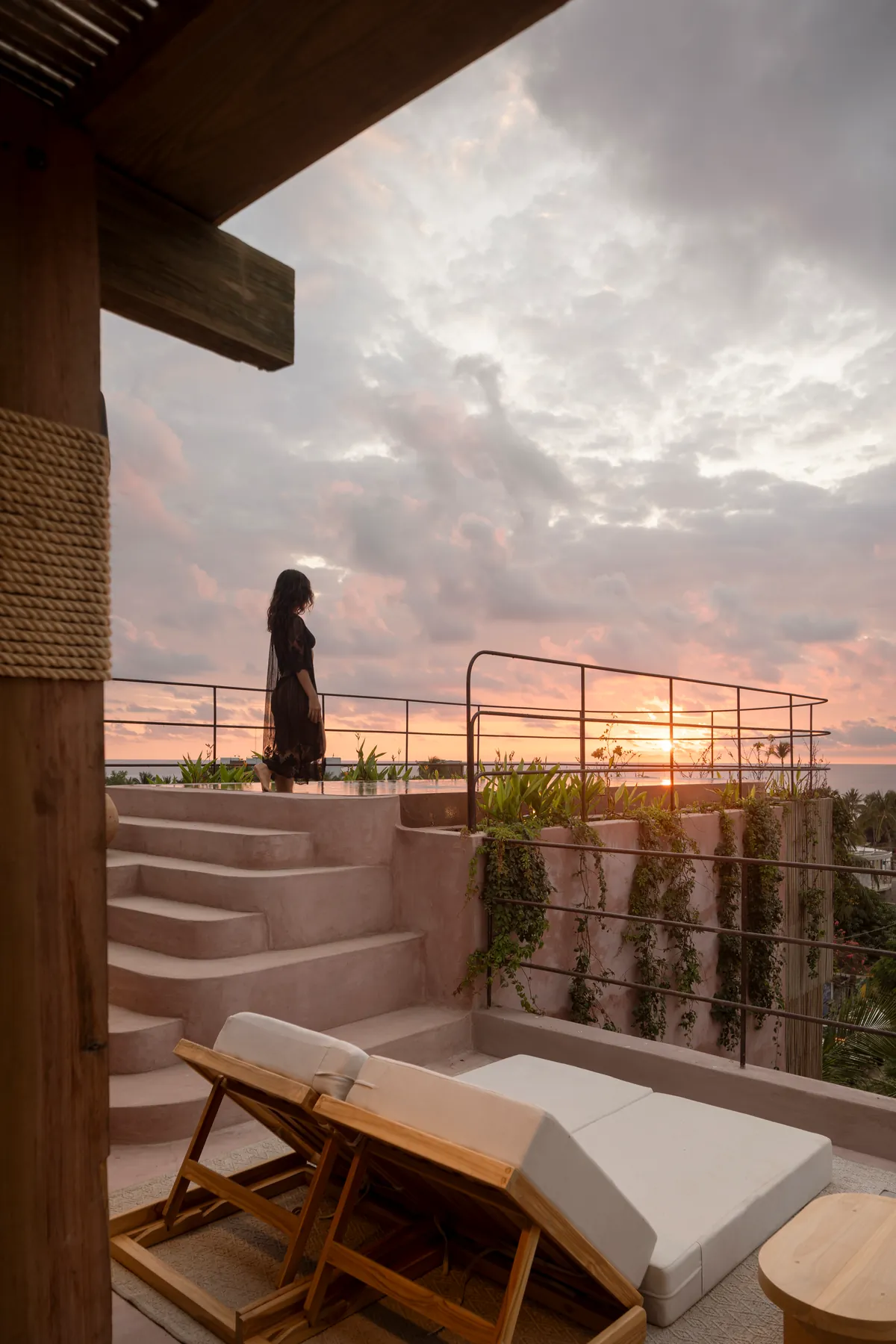 Mujer en lo alto de los escalones de chukum de la azotea al atardecer, con camastros de madera y el sol poniéndose sobre el mar