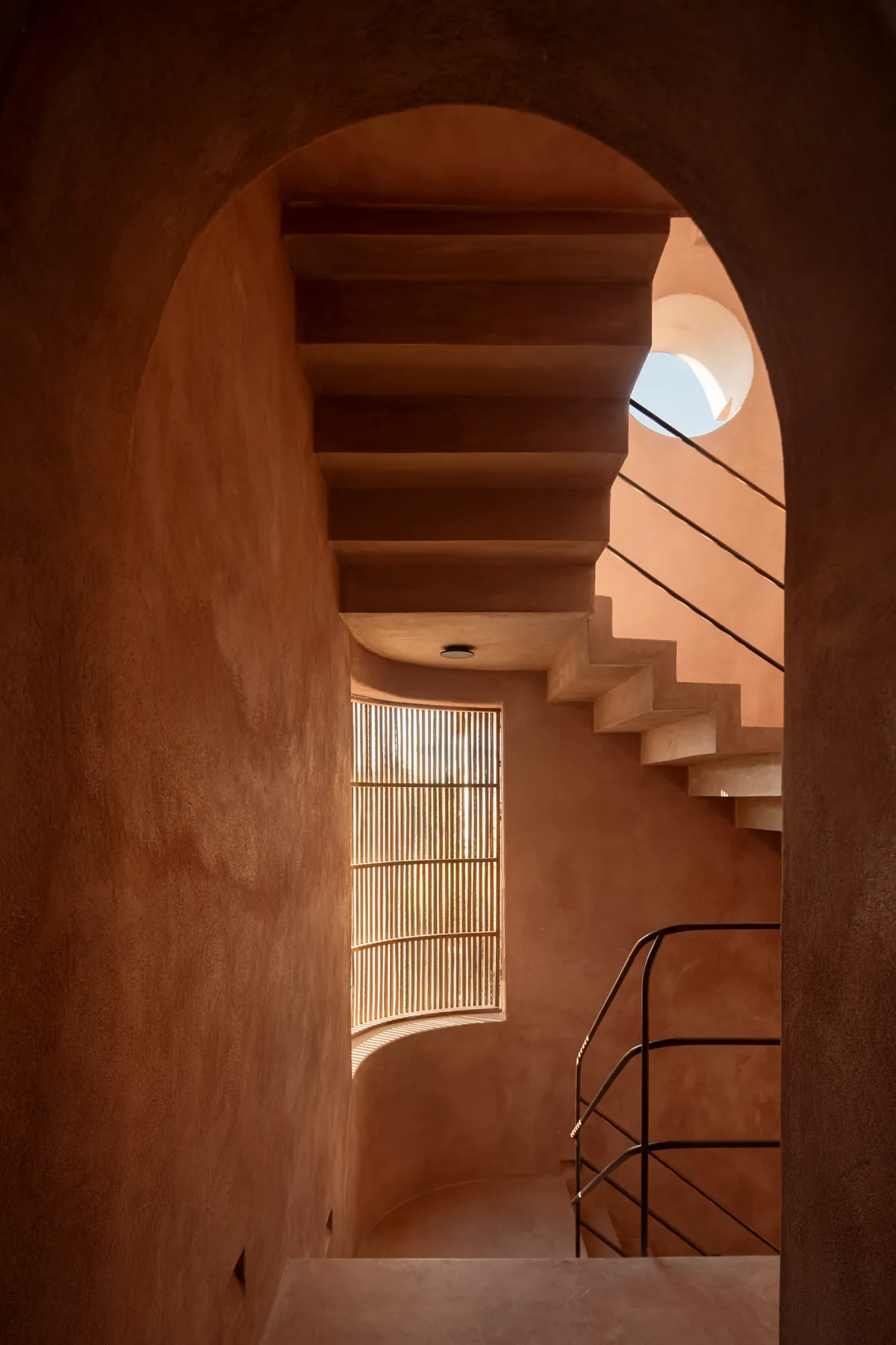 Arco de chukum rosado que enmarca una escalera de concreto, una ventana curva de varas y un óculo de luz