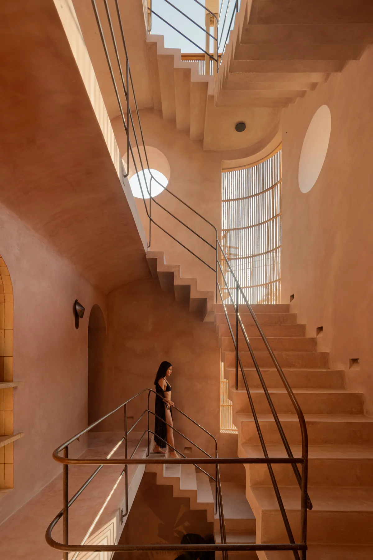 Escalera de chukum bañada de luz, con barandales metálicos, óculos y una ventana curva de varas; una mujer baja los peldaños